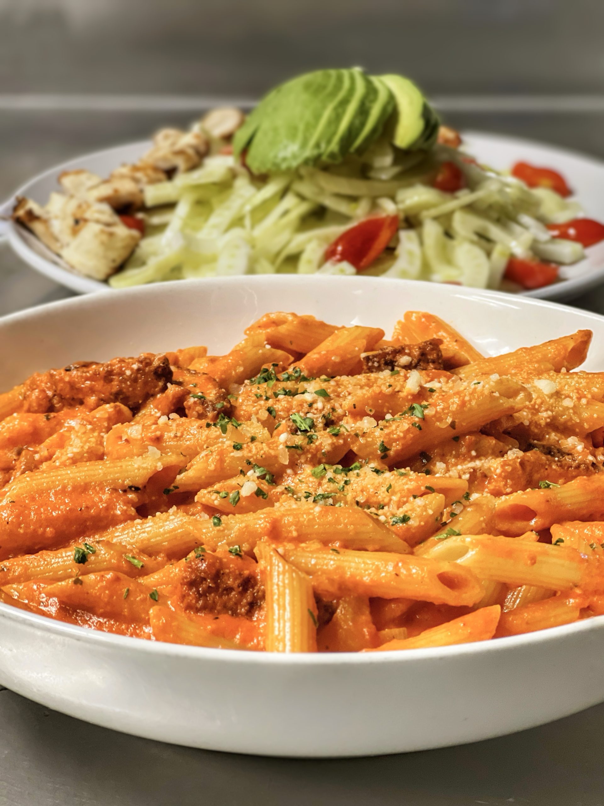 Penne pasta with vodka sauce in focus and grilled chicken cobb salad out of focus